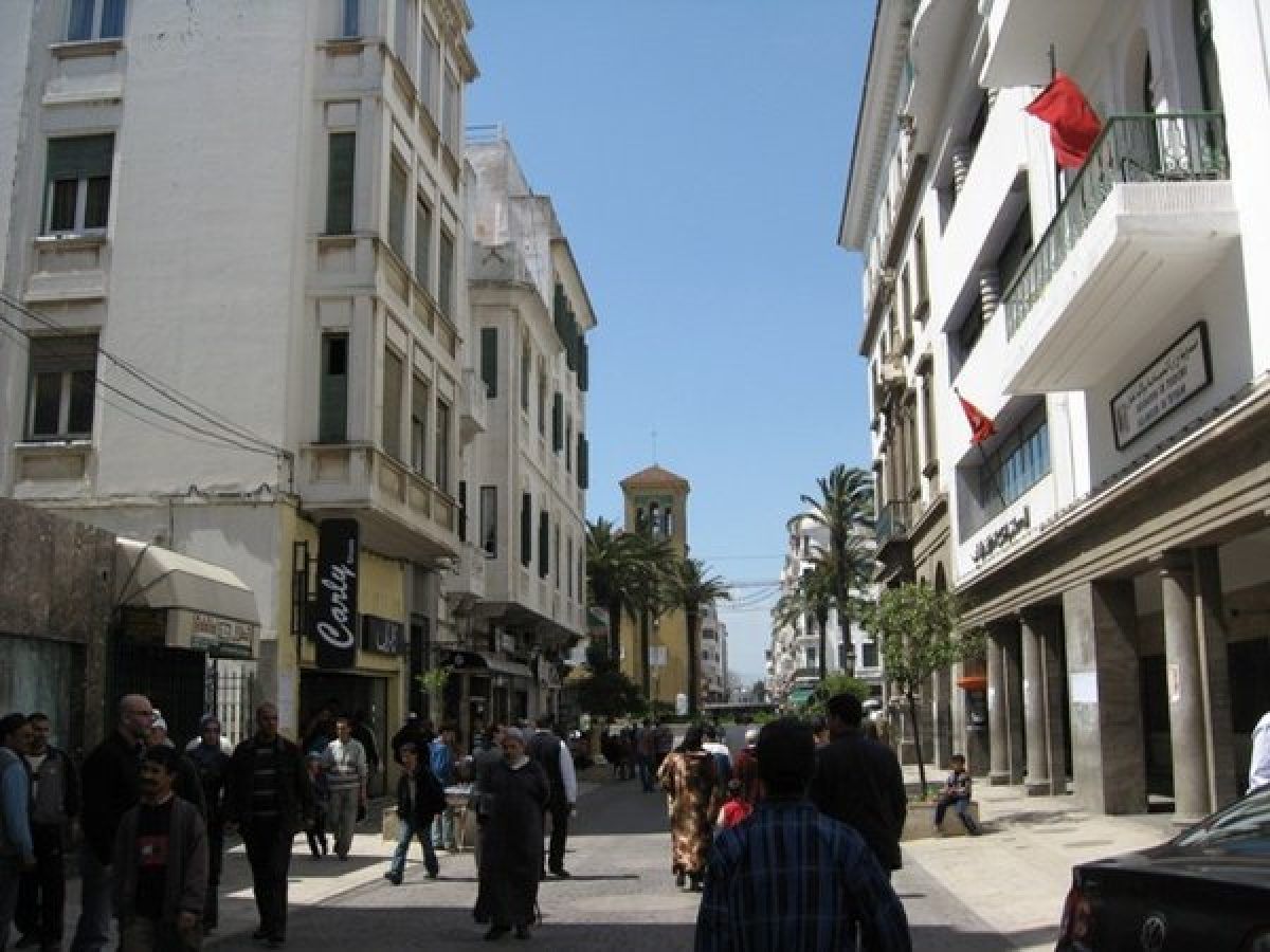 Calle concurrida del ensanche de Tetuán con edificios de estilo español, palmeras y viajeros paseando bajo las banderas de Marruecos