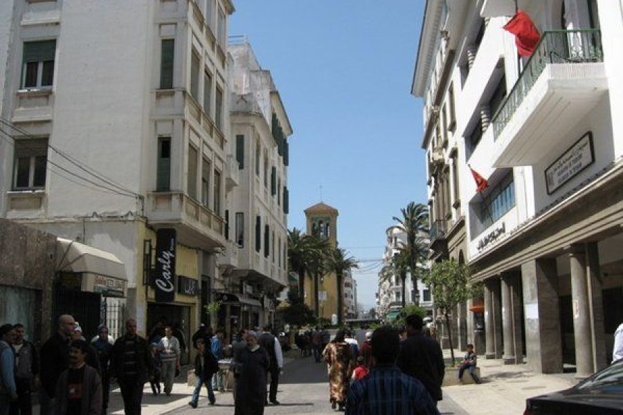 Calle concurrida del ensanche de Tetuán con edificios de estilo español, palmeras y viajeros paseando bajo las banderas de Marruecos
