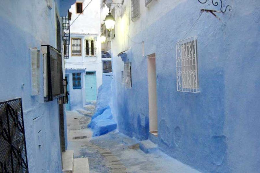 Callejón estrecho de Chefchaouen con paredes azules, escalones de piedra y farola encendida al atardecer en la ciudad azul