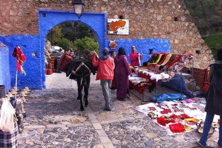 Puerta de arco azul de Chefchaouen con burro cargado y viajeros entre puestos de artesanía y mantas coloridas