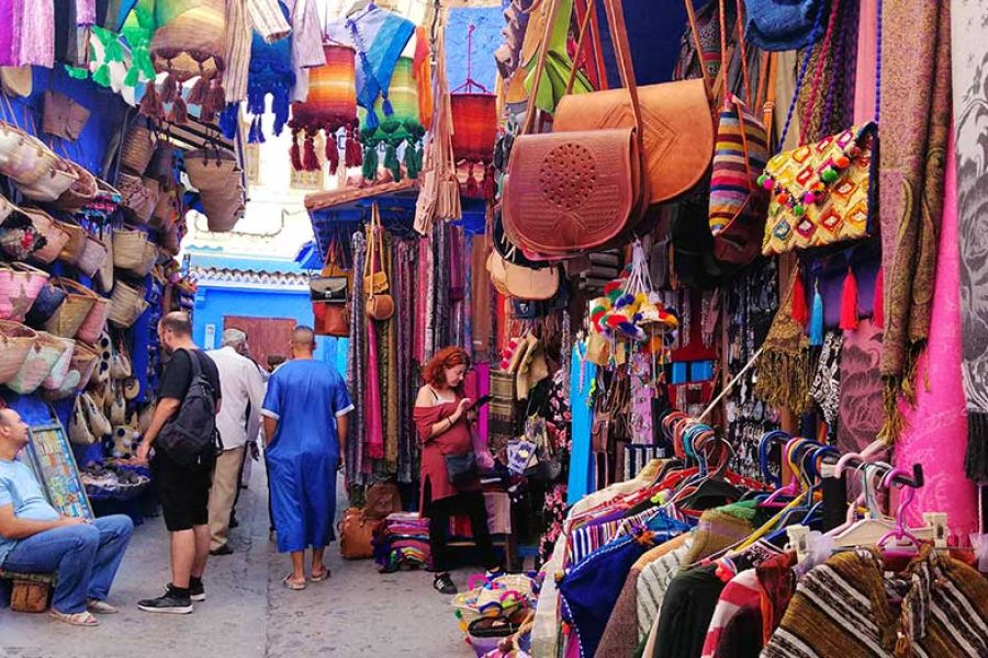 Viajeros comprando en concurrida callejuela de artesanía de Chefchaouen con bolsos, cestas y telas de colores