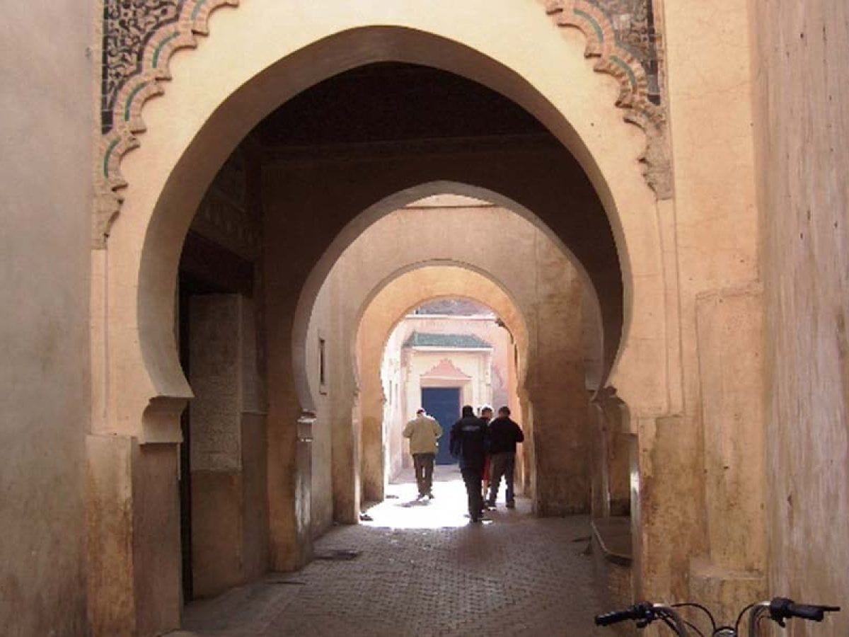 Corredor de arcos de herradura en un edificio histórico de la medina de Marrakech con viajeros caminando hacia la luz al fondo