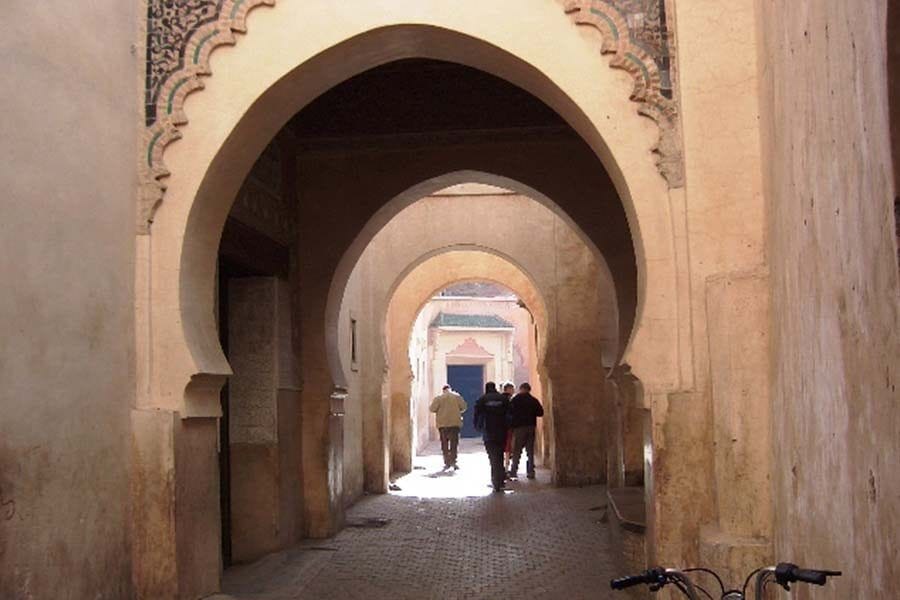 Corredor de arcos de herradura en un edificio histórico de la medina de Marrakech con viajeros caminando hacia la luz al fondo