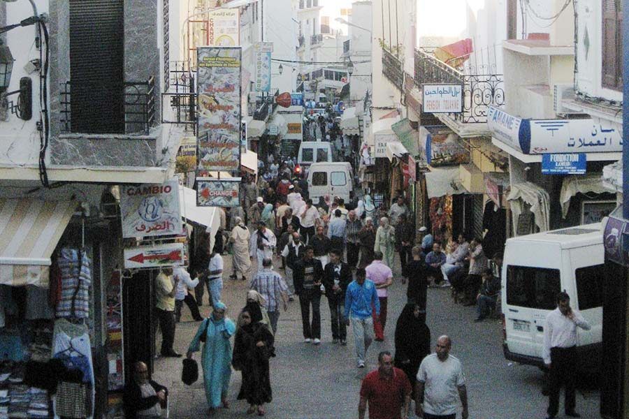 Calle comercial concurrida de la medina de Tánger con viajeros, tiendas y vehículos en primer plano