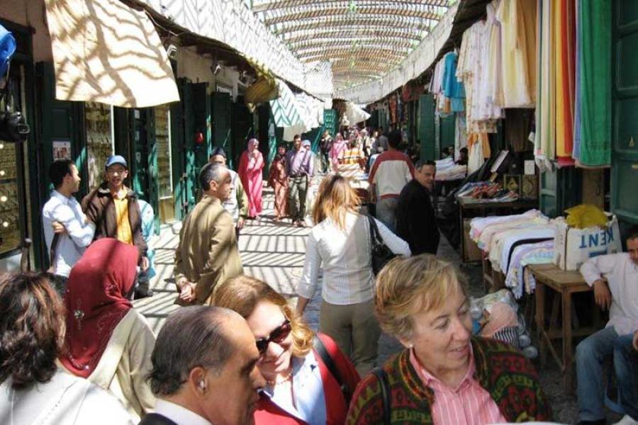 Viajeras y viajeros paseando por el mercado cubierto de la medina de Tetuán con tiendas de ropa y tejidos