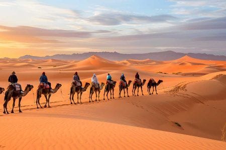 Caravana de viajeros montados en camellos cruzando las dunas doradas del desierto de Merzouga al atardecer con el sol en el horizonte
