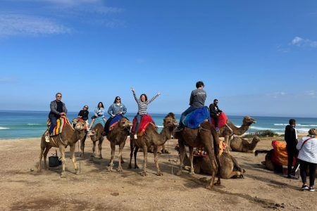 Jóvenes viajeros montando camellos en la playa del norte de Marruecos con el Atlántico al fondo
