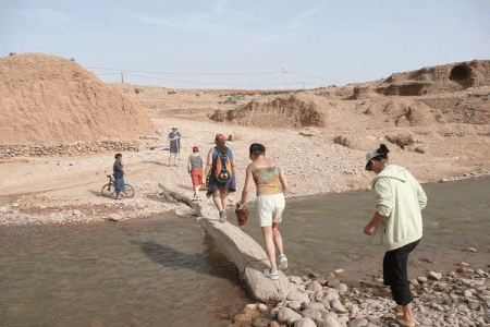 Jóvenes viajeros cruzando un río pedregoso en el paisaje árido del valle del Dadés con colinas ocres al fondo