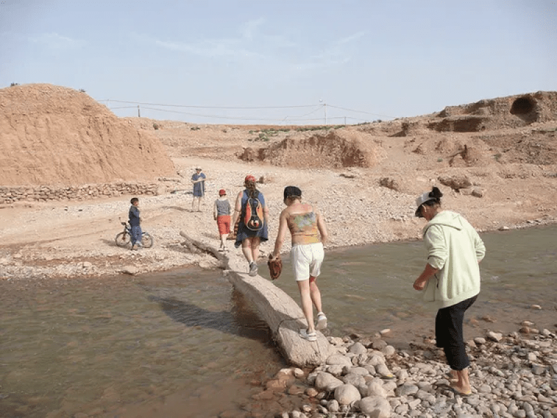 Jóvenes viajeros cruzando un río pedregoso en el paisaje árido del valle del Dadés con colinas ocres al fondo