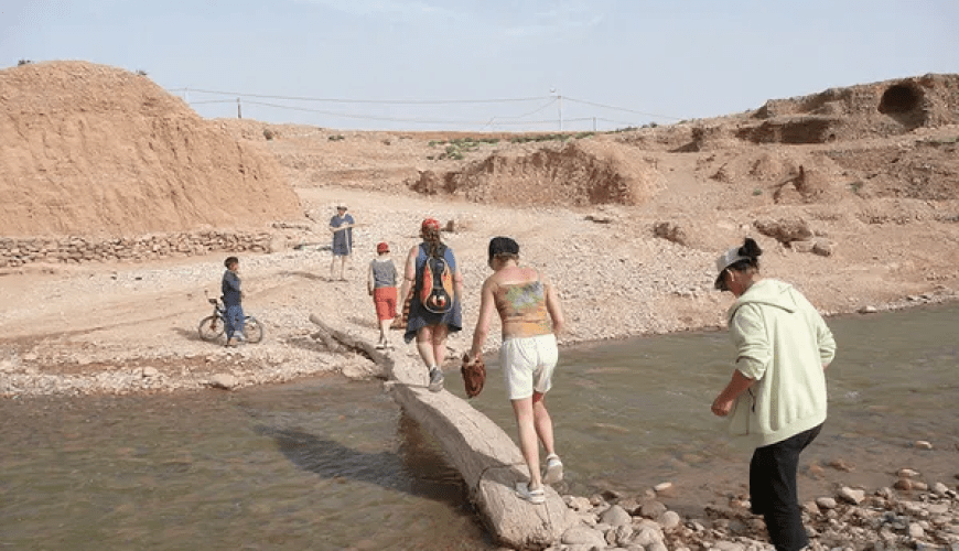 Jóvenes viajeros cruzando un río pedregoso en el paisaje árido del valle del Dadés con colinas ocres al fondo