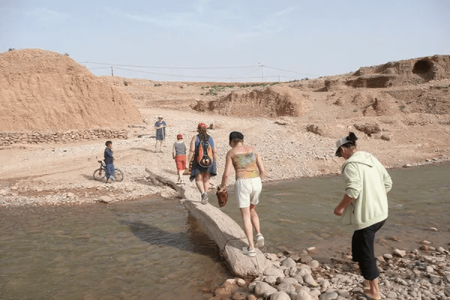 Jóvenes viajeros cruzando un río pedregoso en el paisaje árido del valle del Dadés con colinas ocres al fondo