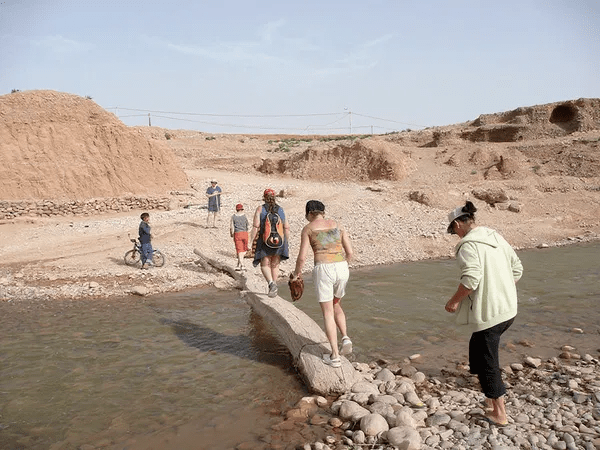 Jóvenes viajeros cruzando un río pedregoso en el paisaje árido del valle del Dadés con colinas ocres al fondo