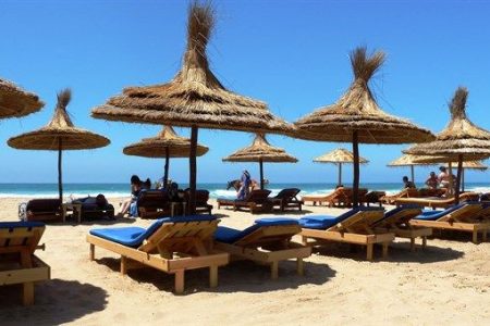 Playa de Asilah en verano con tumbonas de madera y sombrillas de paja a la orilla del Atlántico bajo un cielo azul despejado