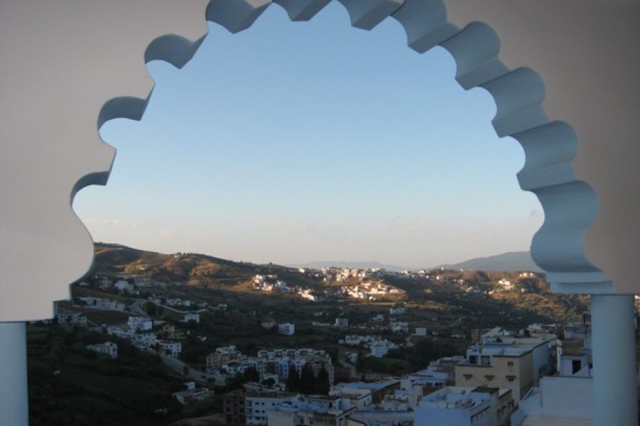 Vista de Chefchaouen al atardecer enmarcada en un arco morisco escalonado del Hotel Parador, con la ciudad blanca y azul extendiéndose por las montañas del Rif