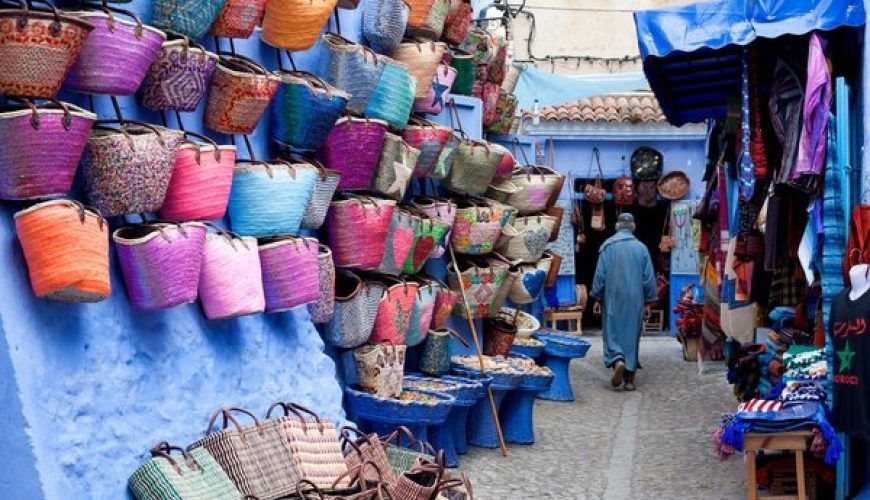 Callejuela de Chefchaouen con cestos y bolsos artesanales de colores colgados en la pared azul y un viajero caminando entre tiendas