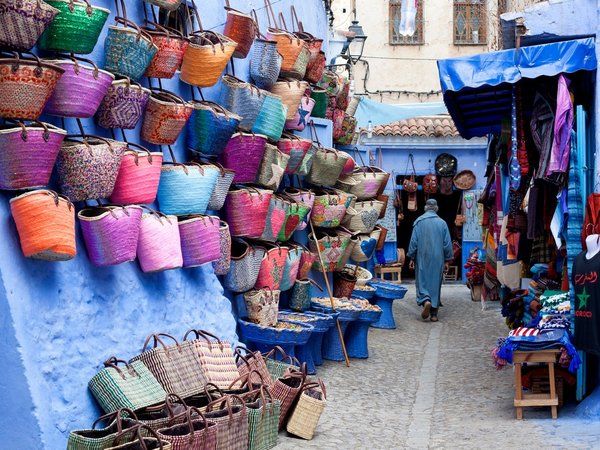 Callejuela de Chefchaouen con cestos y bolsos artesanales de colores colgados en la pared azul y un viajero caminando entre tiendas